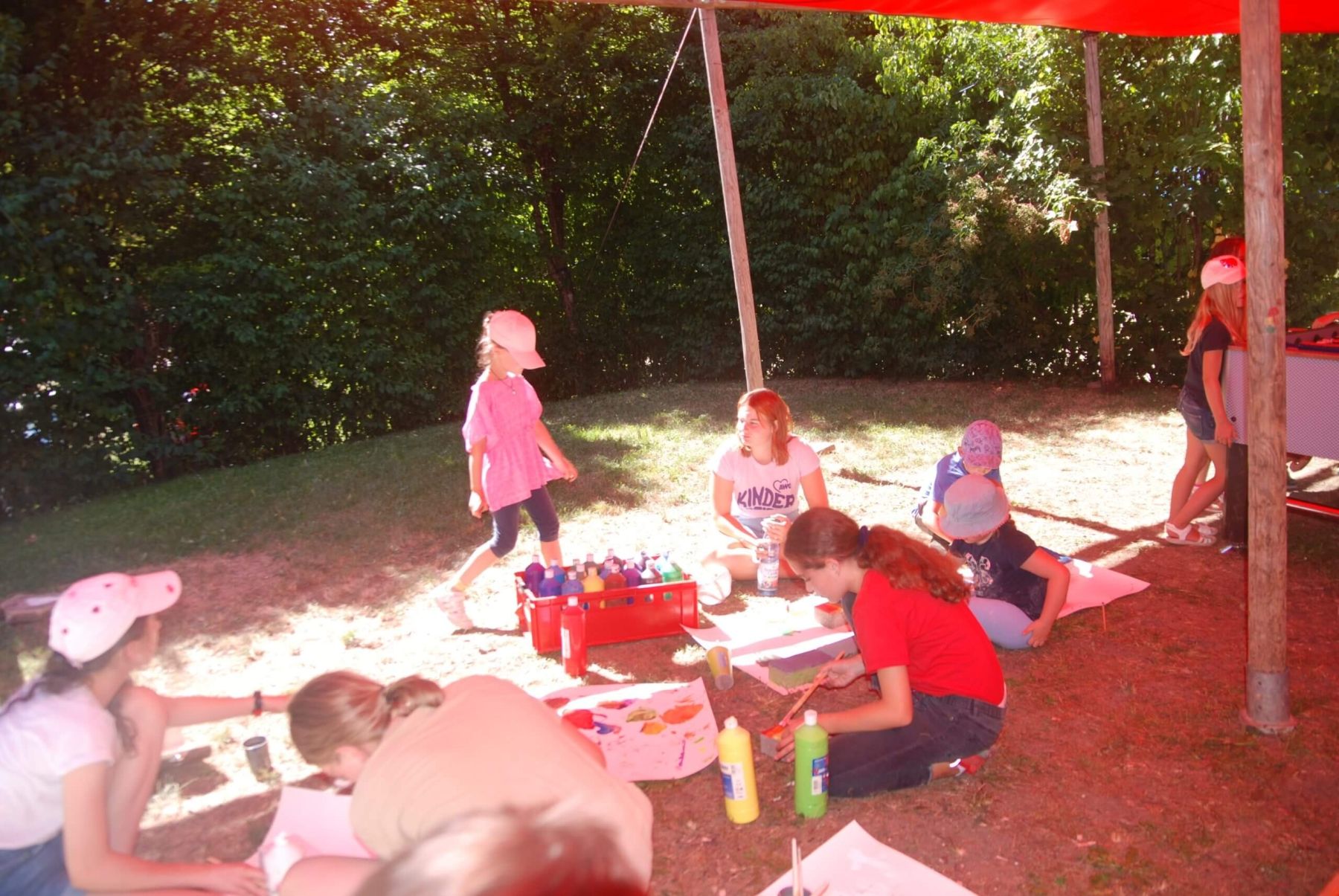 Kinder sitzen unter einem Sonnenschutz und malen mit bunten Farben auf Papier.