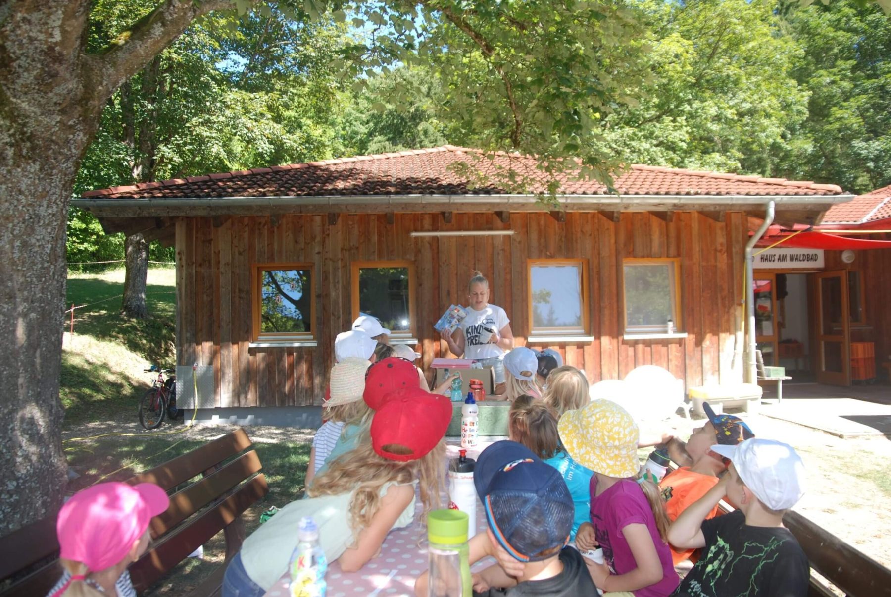 Eine Betreuerin liest einer Gruppe Kinder vor, die an einem Tisch vor einer Holzhütte sitzen.