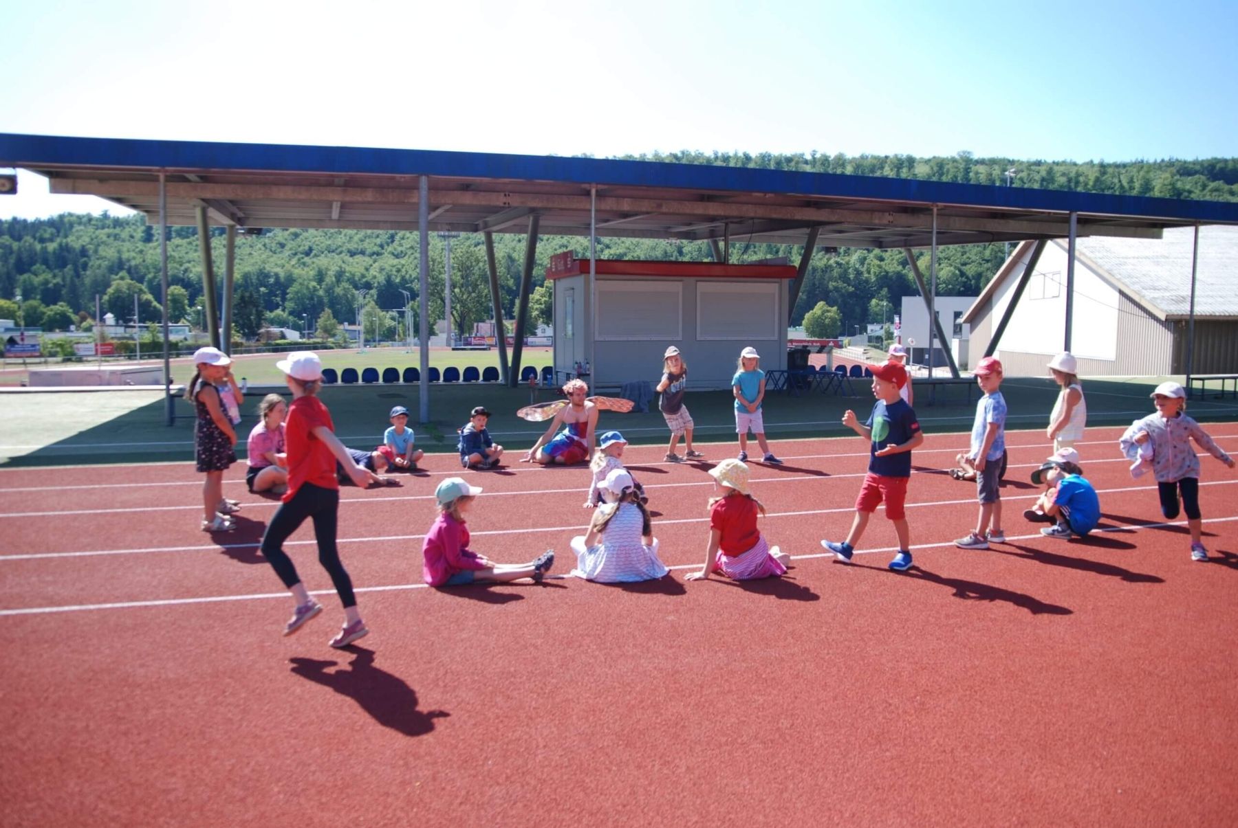 Kinder spielen ein Bewegungsspiel auf einem Sportplatz bei sonnigem Wetter.