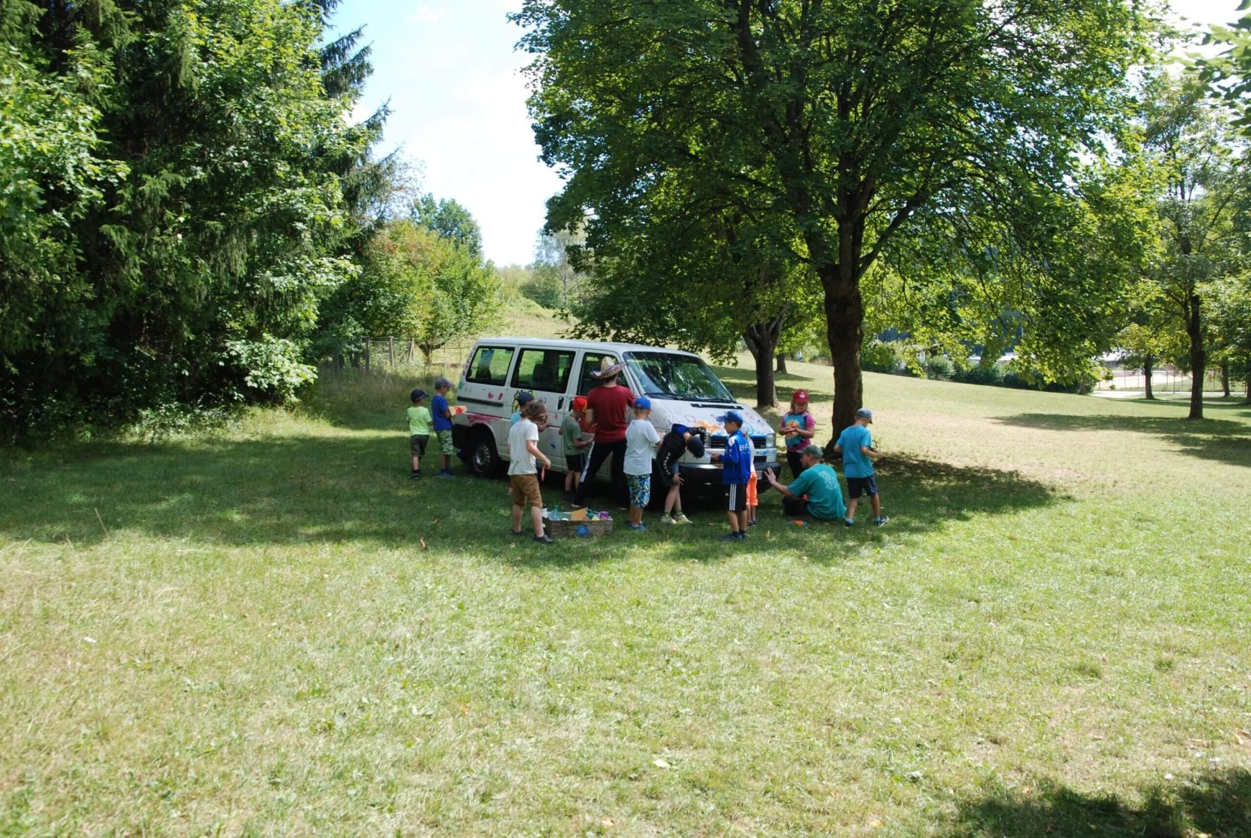 Eine Gruppe Kinder bastelt und spielt rund um ein Fahrzeug auf einer Wiese unter Bäumen.