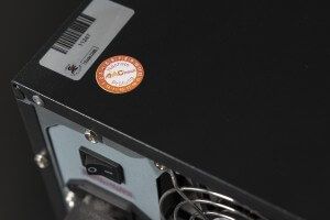 Das Bild zeigt die Rückseite eines elektronischen Geräts mit Netzschalter, Lüftungsgitter und einer Prüfplakette zur Sicherheitskennzeichnung.